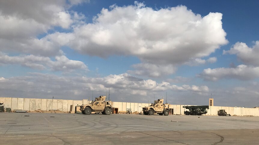 Sandy coloured military vehicles are parked in the distance an an airbase next to a fence with blue skies and clouds behind