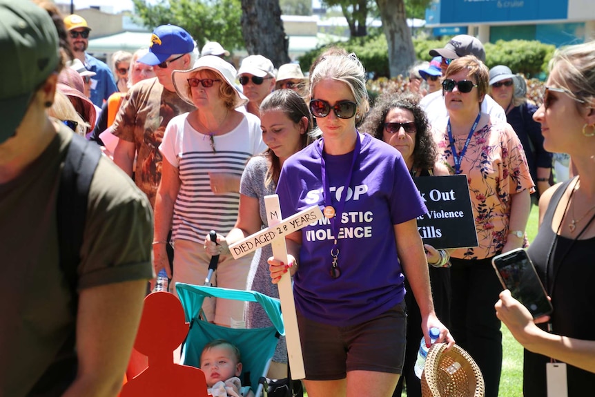 A crowd of men and women including a young woman in a purple shirt holding a white cross walk outdoors during a rally.