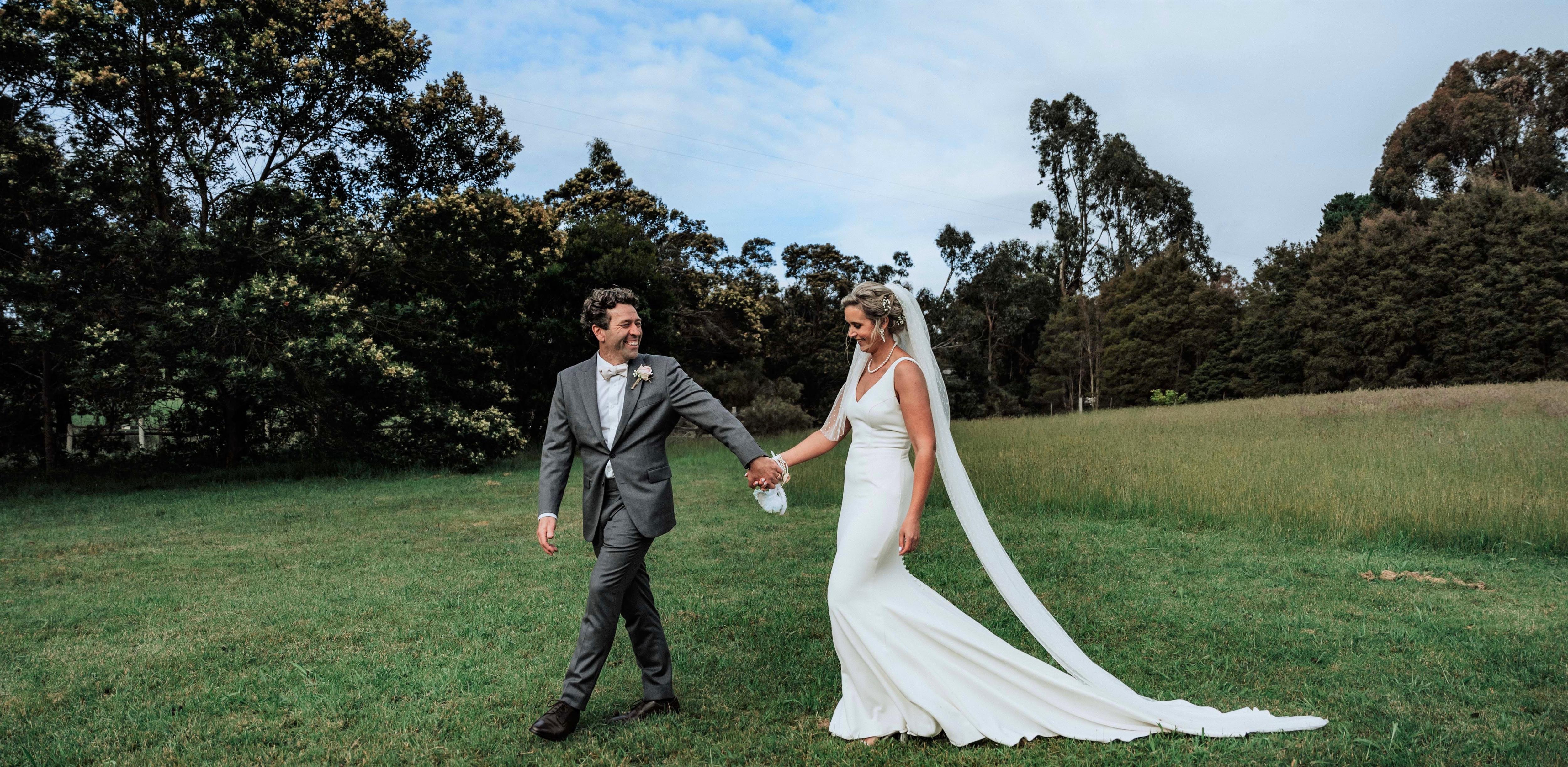 A man in a suit and a woman in a wedding dress holding hands with greenery behind