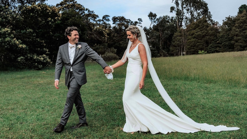 A man in a suit and a woman in a wedding dress holding hands with greenery behind