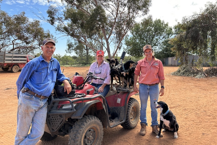 Smiling older man, denim shirt, jeans, cap, leans on red quad bike driven by woman in pink shirt, cap, girl dogs stand beside.