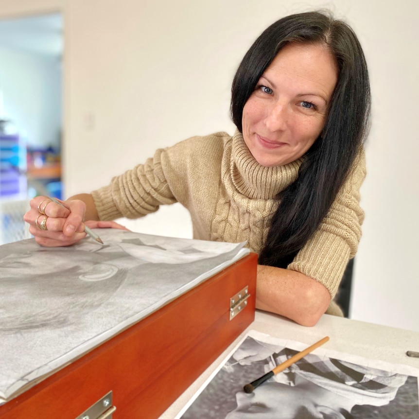 Woman with long, dark hair sits at the end of a kitchen bench sketching. She is smiling.