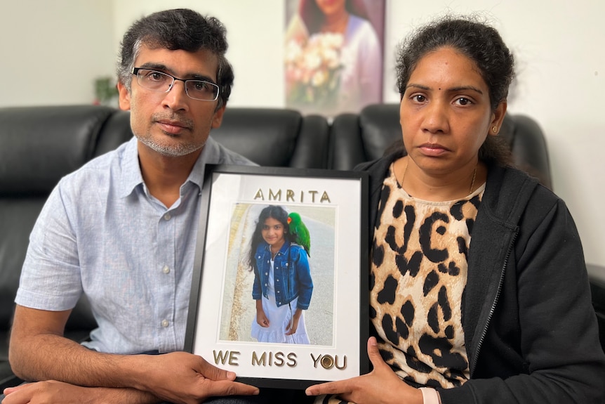 A man and a woman hold up a photo frame with a picture of a girl and the words 'We Miss You Amrita'.