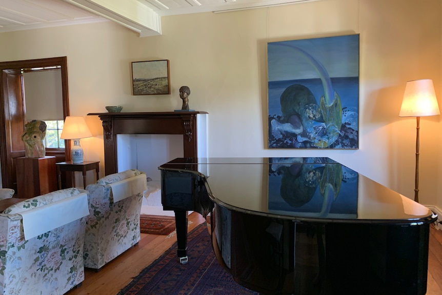 An old-style sitting room with a grand piano in the centre and a backdrop of artworks from abstract to landscape. 