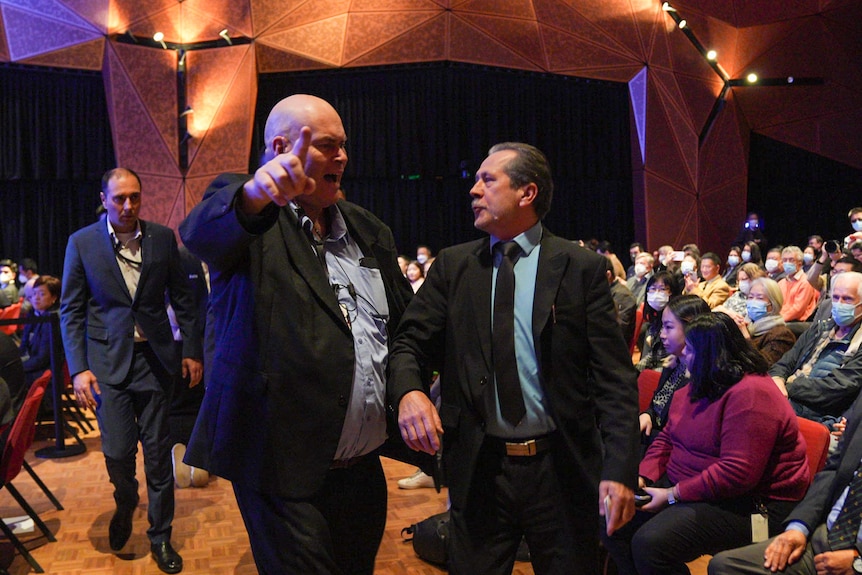 A man points and escorts another man in suit and tie out of a hall