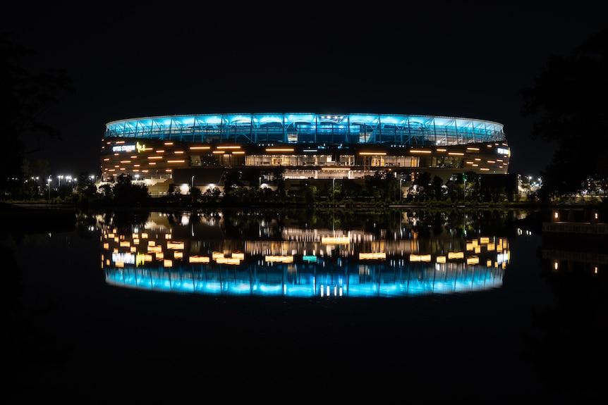 Large building with blue light on rim of roof, reflected in river surface, surrounded by black darkness.
