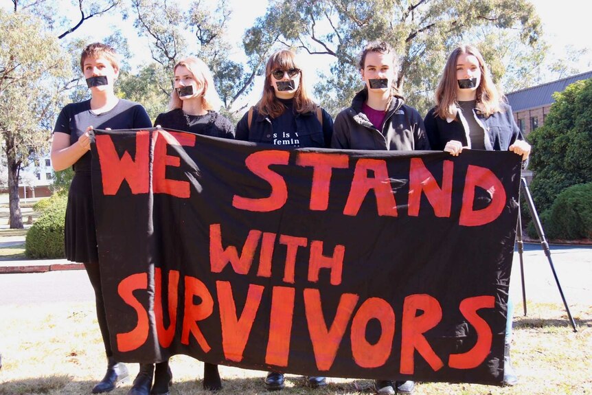 Female students with tape over their mouths hold a sign saying 'we stand with survivors'.