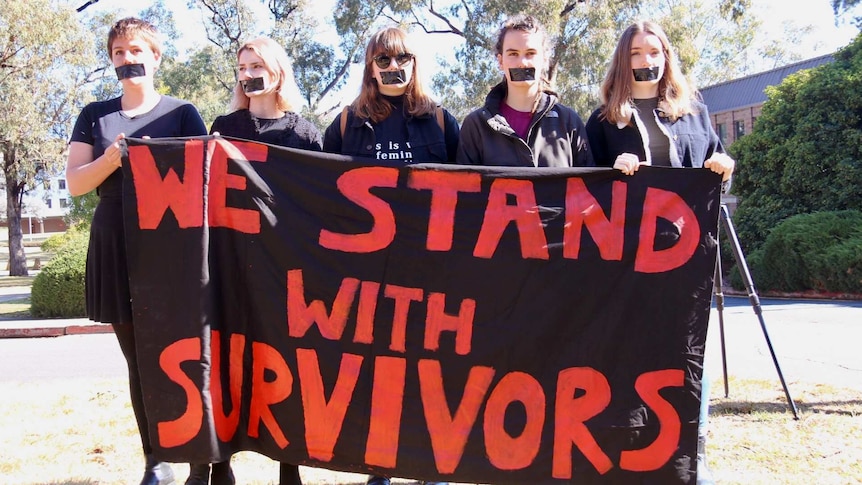 Female students with tape over their mouths hold a sign saying 'we stand with survivors'.