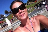 A woman with brown hair tied back and big sunglasses takes a selfie in a park on a sunny day.