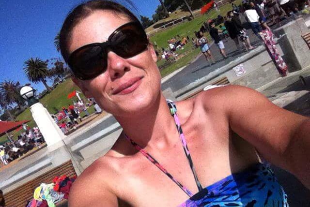 A woman with brown hair tied back and big sunglasses takes a selfie in a park on a sunny day.