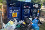 Four charity bins from Lifeline overflowing with donations and items strewn in front of them.