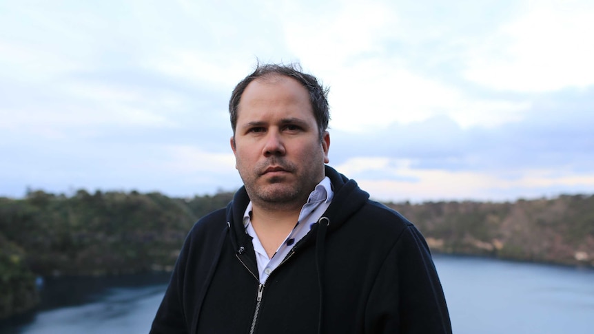 A theatre director with a serious look on his face stands with Mount Gambier's Blue Lake in the background