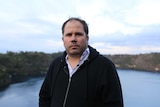 A theatre director with a serious look on his face stands with Mount Gambier's Blue Lake in the background