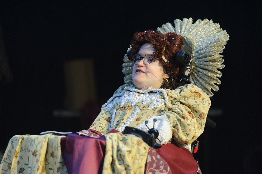 A woman in a wheelchair with a floral dress, a curly wig and a large ruff stuck to the back of her head rest