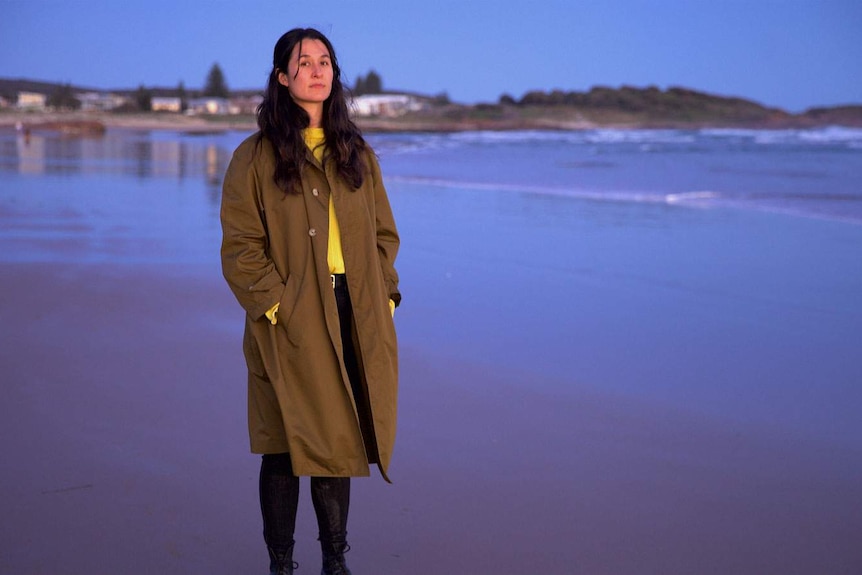 The isnger, with long brown hair and wearing an over-sized trench coat stands on a beach at low tide.