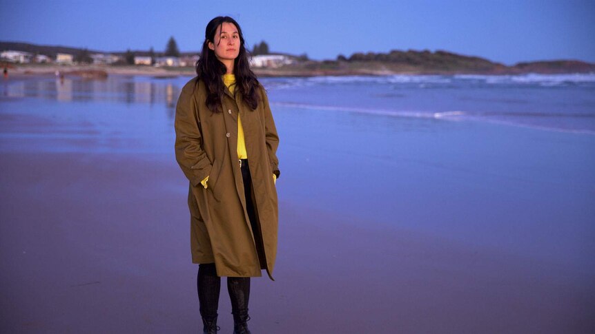 The isnger, with long brown hair and wearing an over-sized trench coat stands on a beach at low tide.