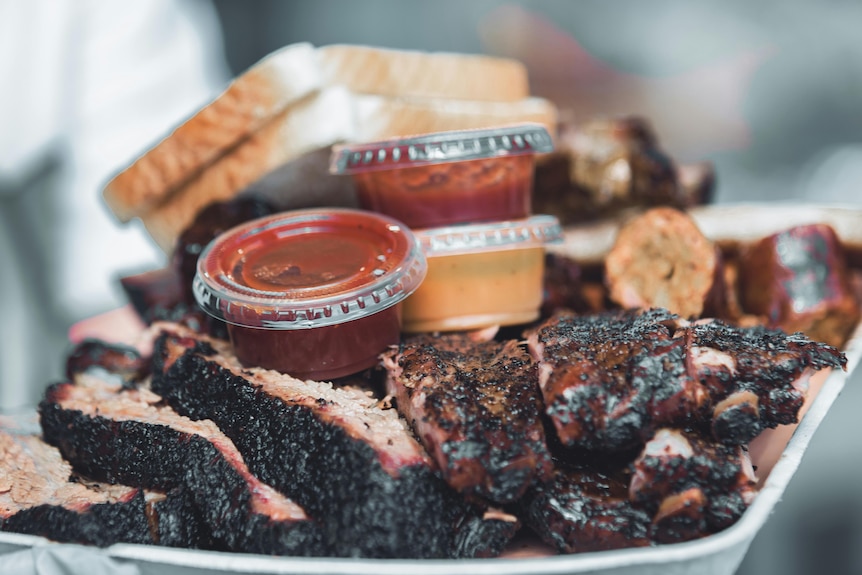 Thick slices of barbecued meat piled on a plate, alongside slices of bread and containers of red and brown sauce.