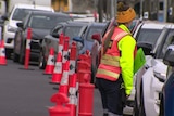 A row of cars wait queue next to traffic cones.