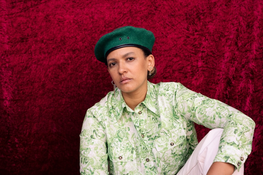 A woman sits with a hand draped on her knee in front of a deep maroon velvet backdrop. She wears a green beret & printed shirt.