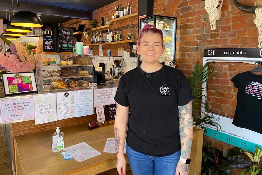 A woman in a black shirt with pink hair smiles while standing in front of a café counter.