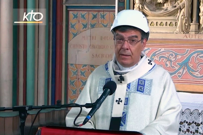 The archbishop standing at a pulpit with a microphone wears a hard hat