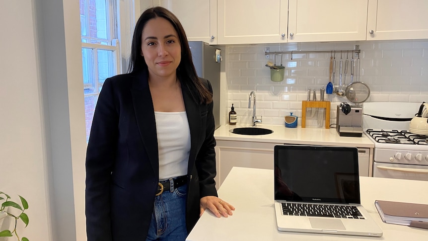 A woman wearing a white top and a black blazer stands next to a benchtop with a laptop on it.