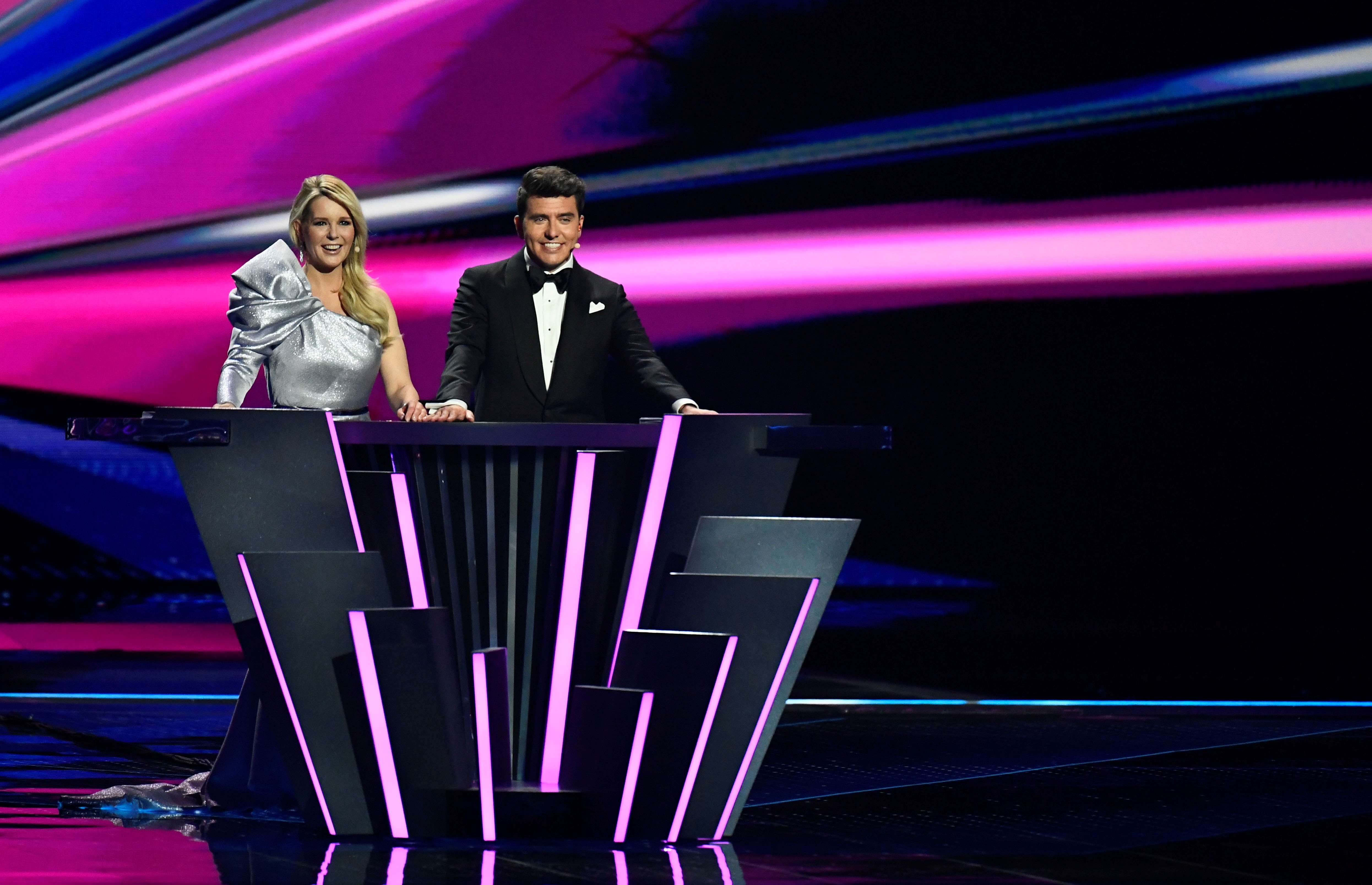 A blonde woman stands next to a brunette man. She wears a silver dress and he wears a black suit. They're at a podium.