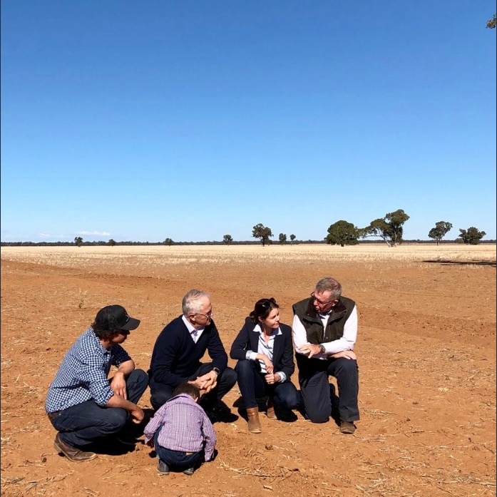 A group of people including Malcolm Turnbull kneel in the brown dirt. The sky is blue and a few trees are visible on the horizon