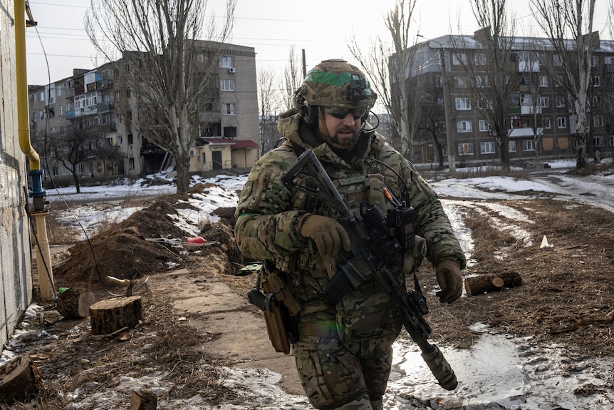 A soldier in battle fatigues patrols an unpaved street near residential buildings.