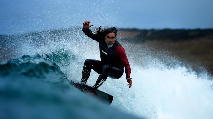 An older man with long hair and a long beard, wearing a full-body wetsuit surfs a wave.