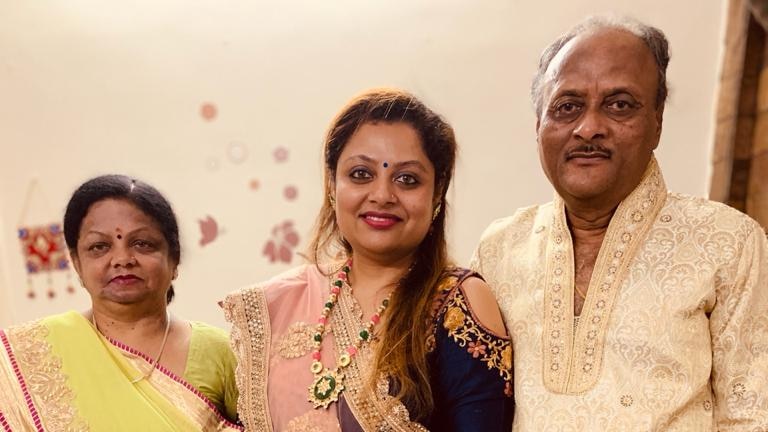 A woman wearing a pink sari stands in the middle of an older woman wearing a yellow sari and a man.