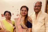 A woman wearing a pink sari stands in the middle of an older woman wearing a yellow sari and a man.