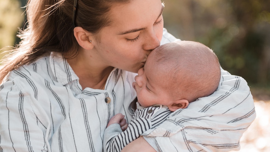 A mother in white shirt with brown long hair kisses her baby on his head as she holds him