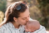 A mother in white shirt with brown long hair kisses her baby on his head as she holds him