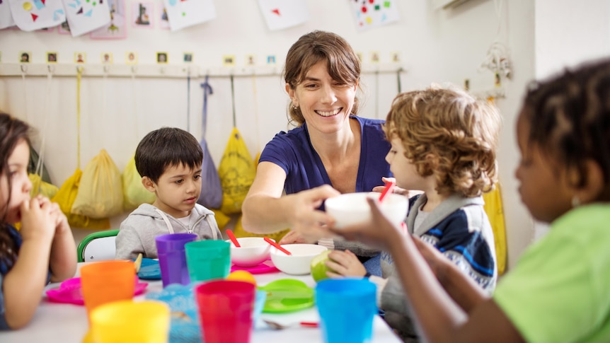 A childcare worker passing bowls to children seated at a table in a kindergarten