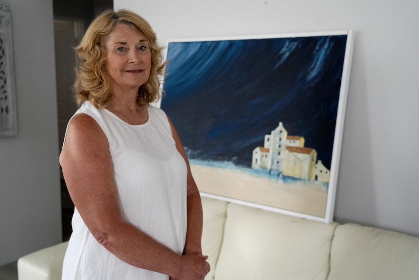 A woman in a white dress stands in front of a large painting of some buildings against a dark blue sky, hands clasped