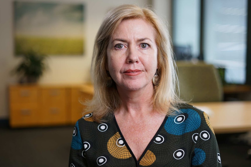 A woman stands in what looks like an office and looks at the camera. She wears a top with geometric patterns and earrings.