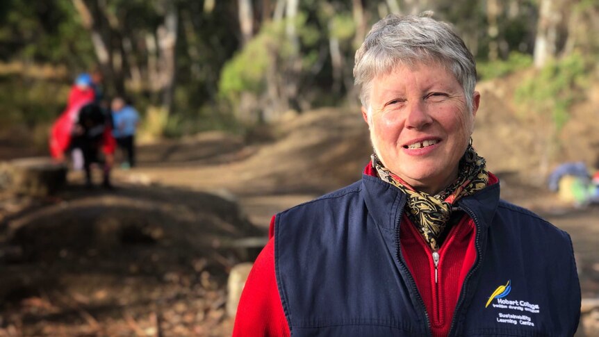 A woman with short grey hair, wearing a blue vest over a red long-sleeved top stands in a bush setting.