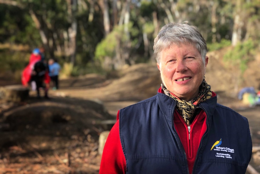 A woman with short grey hair, wearing a blue vest over a red long-sleeved top stands in a bush setting.