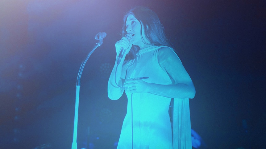 Weyes Blood singing live onstage bathed in blue light and wearing a white caped gown