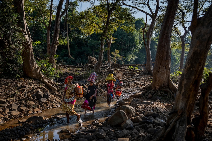 Women carrying bags on top of their heads walk through a river in the middle of a rocky forest. 