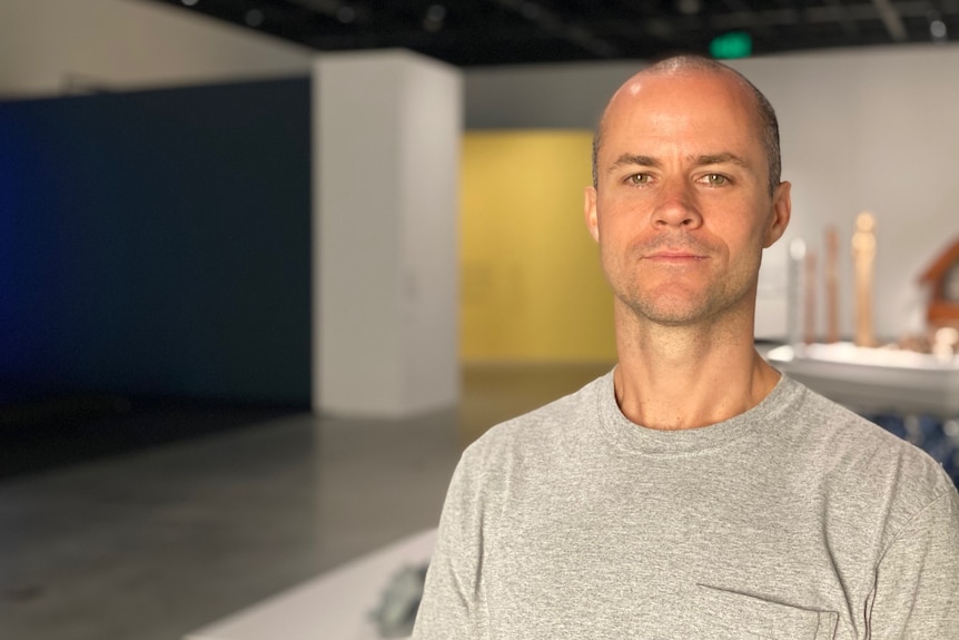 Man with a bald head wearing a t-shirt looking at the camera in a large art gallery space