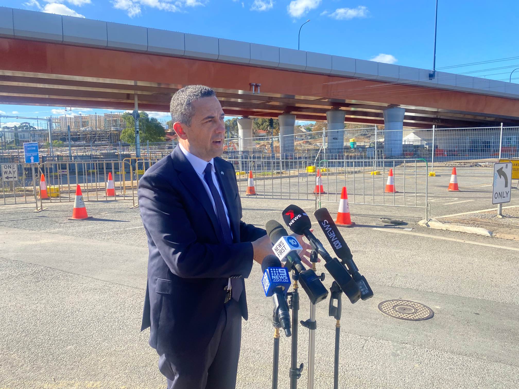 A man in a suit standing behind microphones with a road overpass behind him