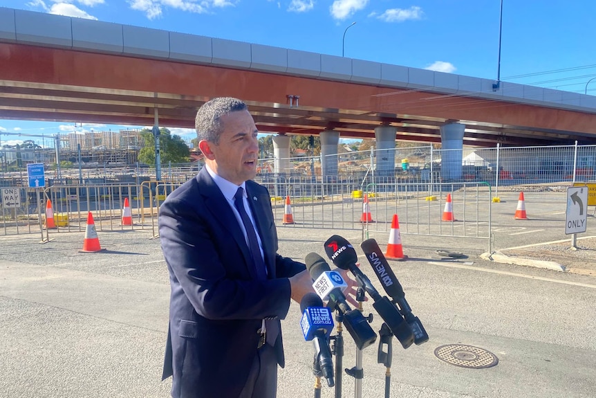 A man in a suit standing behind microphones with a road overpass behind him