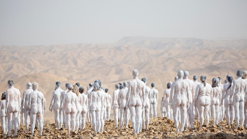 Naked people painted white stand in the desert.