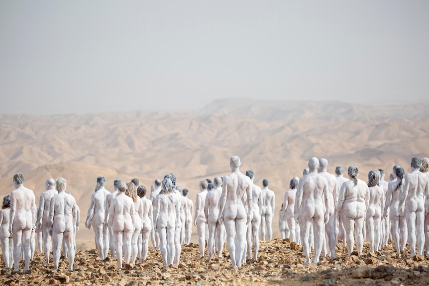 Naked people painted white stand in the desert.