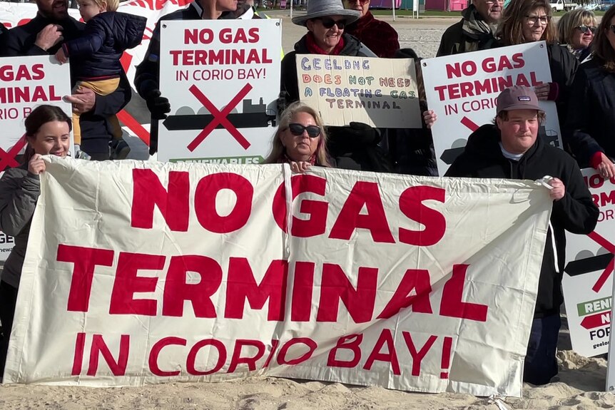 Protesters kneel on the beach holding a sign reading 'NO GAS TERMINAL IN CORIO BAY!' in red text.