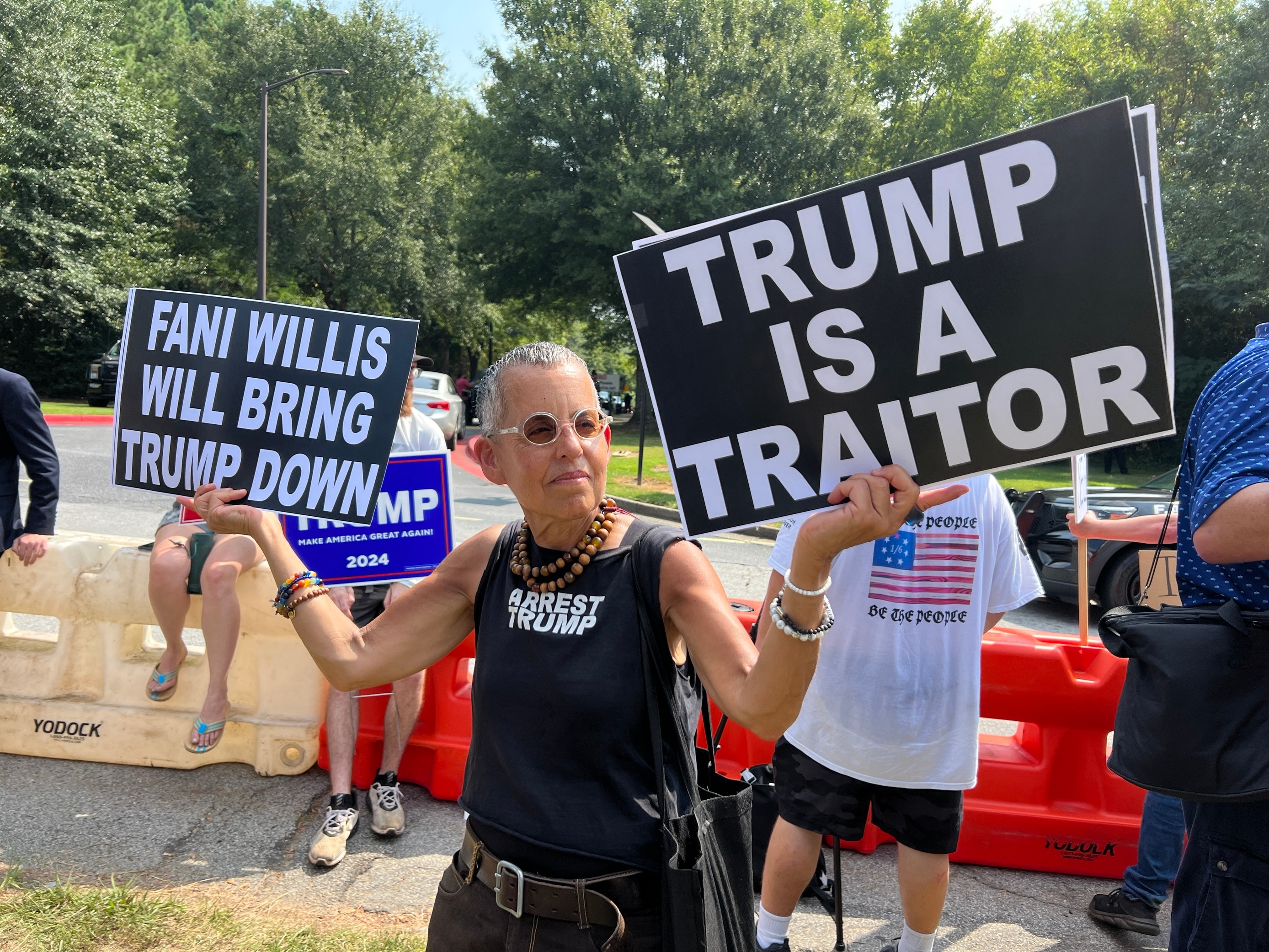 A man in a black T-shirt holds signs saying "Trump is a traitor" and "Fani Willis will bring Trump down"