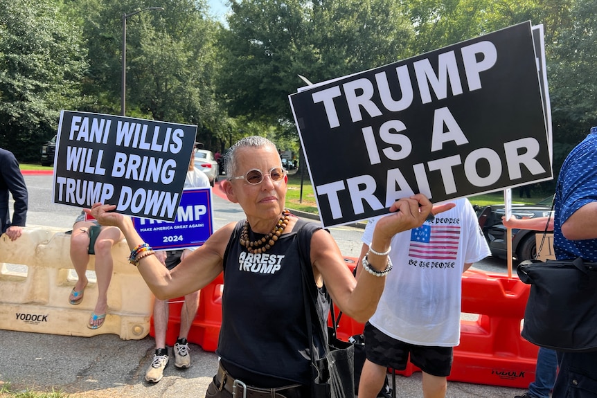 A man in a black T-shirt holds signs saying "Trump is a traitor" and "Fani Willis will bring Trump down"