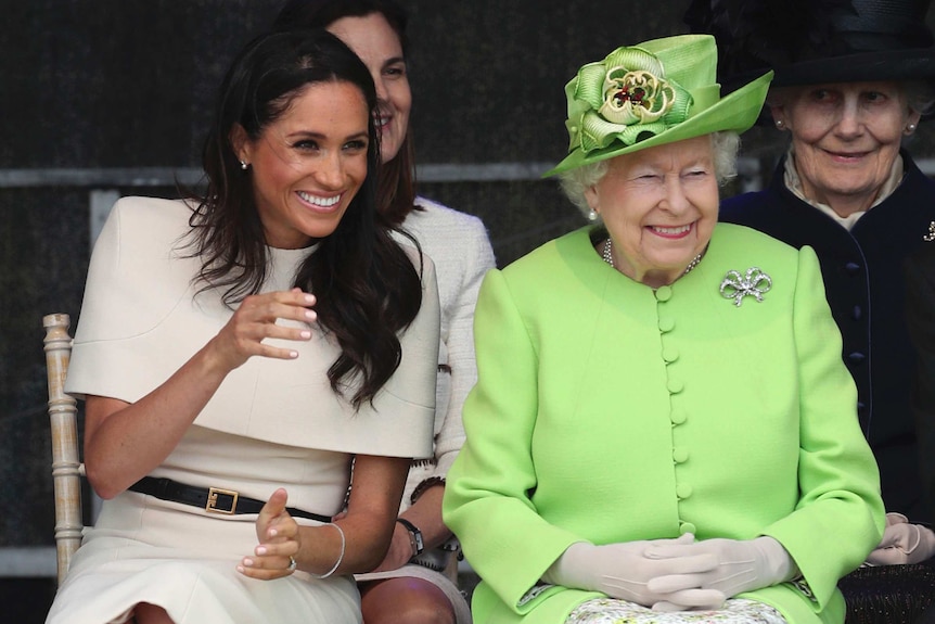 The Duchess of Sussex, sitting next to the Queen, laughs while the Queen smiles.